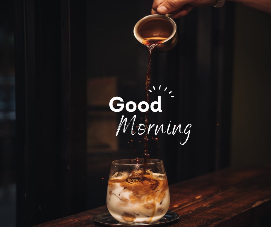 A hand pours coffee into a glass of iced milk on a wooden table, set against a dark background. The text overlay reads, "Good Morning." This image is a stylish example of good morning coffee images.